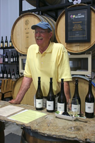 Man with baseball cap in yellow polo shirt standing in front of wine barrels with five bottles of wine arrayed in front of him