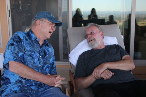 Two men, one on right with gray beard and black shirt and one on left with blue Hawaiian motif shirt and blue baseball cap, chat