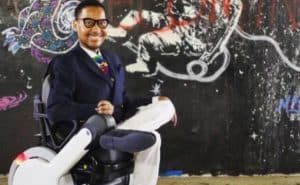 Eddie Ndopu shown sitting in power wheelchair, with white slack and a blue blazer, one foot resting on his knee in front of a wall spray painted with art of an astronaut