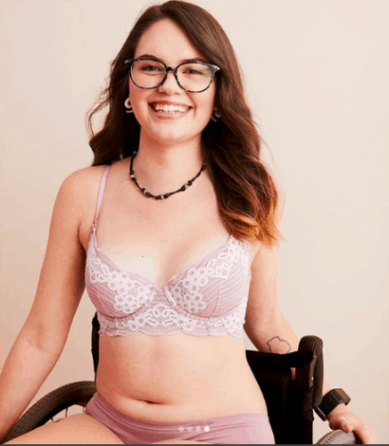 Woman in glasses with brown hair, smiling, sitting in a wheelchair wearing a pink underwear set for inclusive advertising campaign.