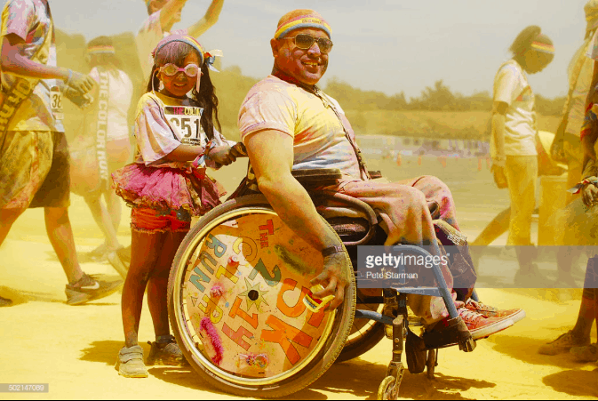 Man in sunglass, dusted in paint with colorful spoke guards on his wheelchair. His young daughter by his side, part of the disability collection, sign up open now.