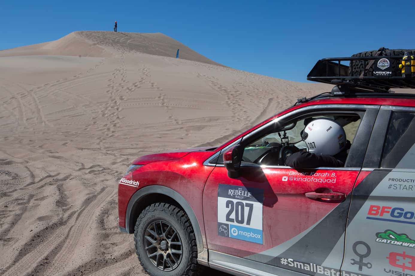 Karah Behrend in white helmet shown in the driver's seat of a red SUV, spotting a line up a sand dune