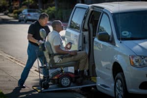 Two men, one in a power wheelchair and another assisting him up a van ramp for wheelchair accessible vehicle service.