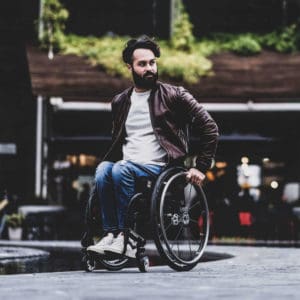 Male wheelchair user with black hair, wearing leather jacket shown pushing Quickie manual wheelchair the Nirtum