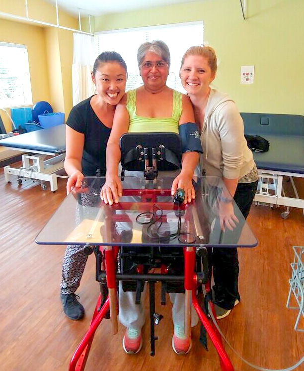 Shubha Ratnaparkhi in standing frame at rehab with two therapists