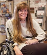Kim Anderson-Erisman, PhD, a white woman with long, light brown hair, sits strapped into her manual wheelchair, wearing a light shirt with striped long-sleeves and documents hanging on the wall behind her.