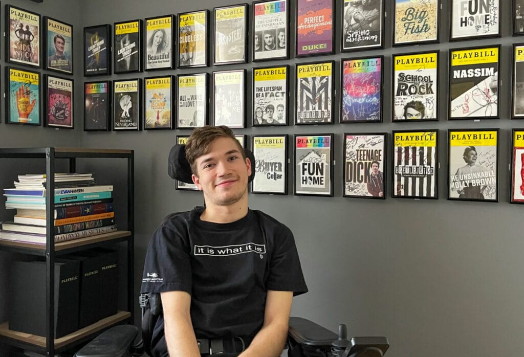 young male in powerchair with wall of framed playbills in background 