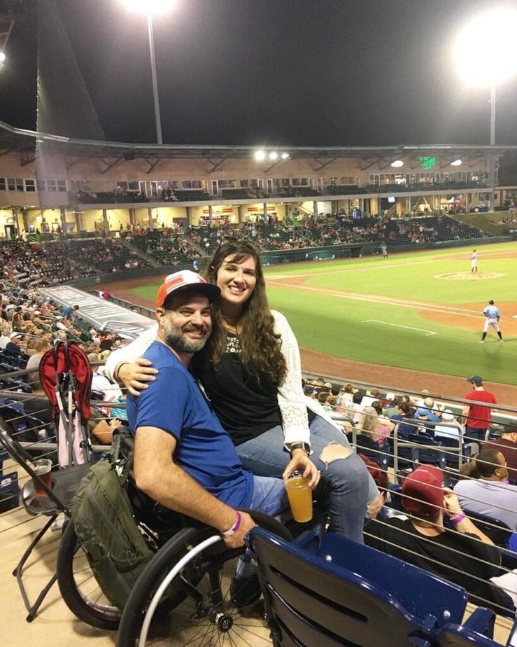 woman sitting on man in wheelchair's lap in the stands at a baseball game