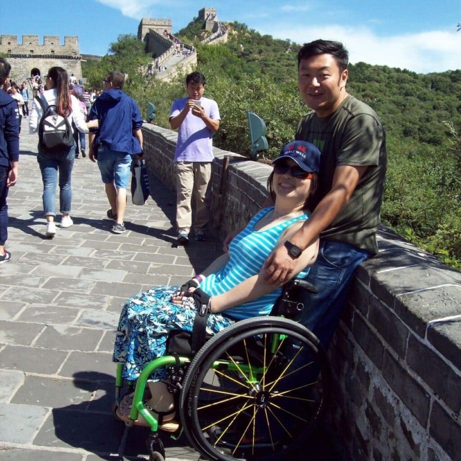 Woman in manual wheelchair sitting on great wall of China with man standing behind her, holdig her shoulders. Both are smiling.