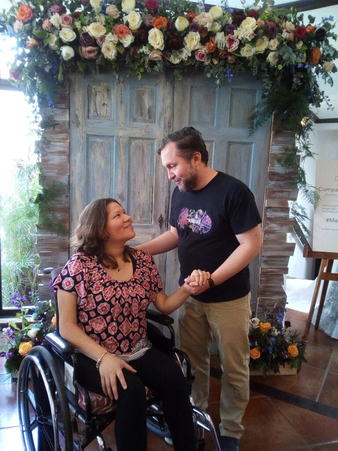 man standing next to wonman in wheelchair holding her hand ond looking each other in the eyes