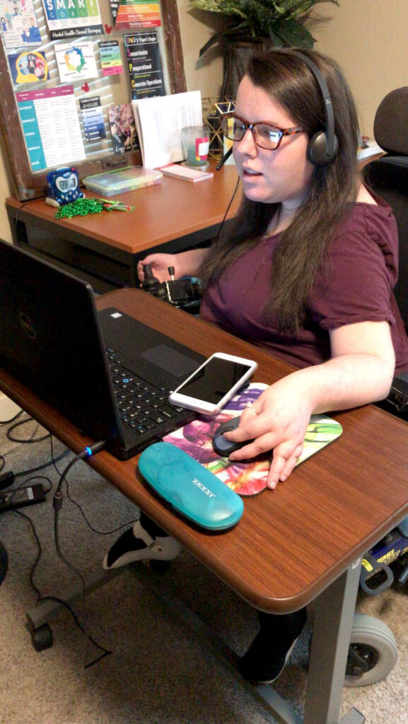 Shows Lauren Presutti, a white woman with long brown hair who sits in a power wheelchair working at a laptop on a height-adjustable desk. She is wearing headphones.