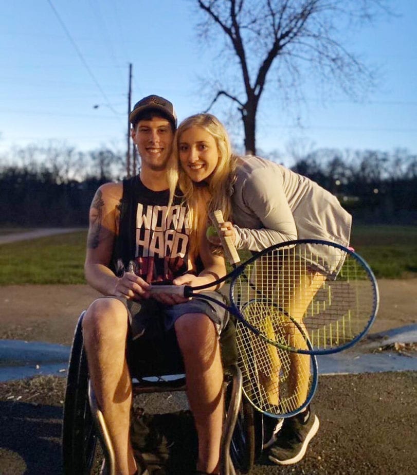 woman crouched down next to man in wheelchair both holding tennis rackets