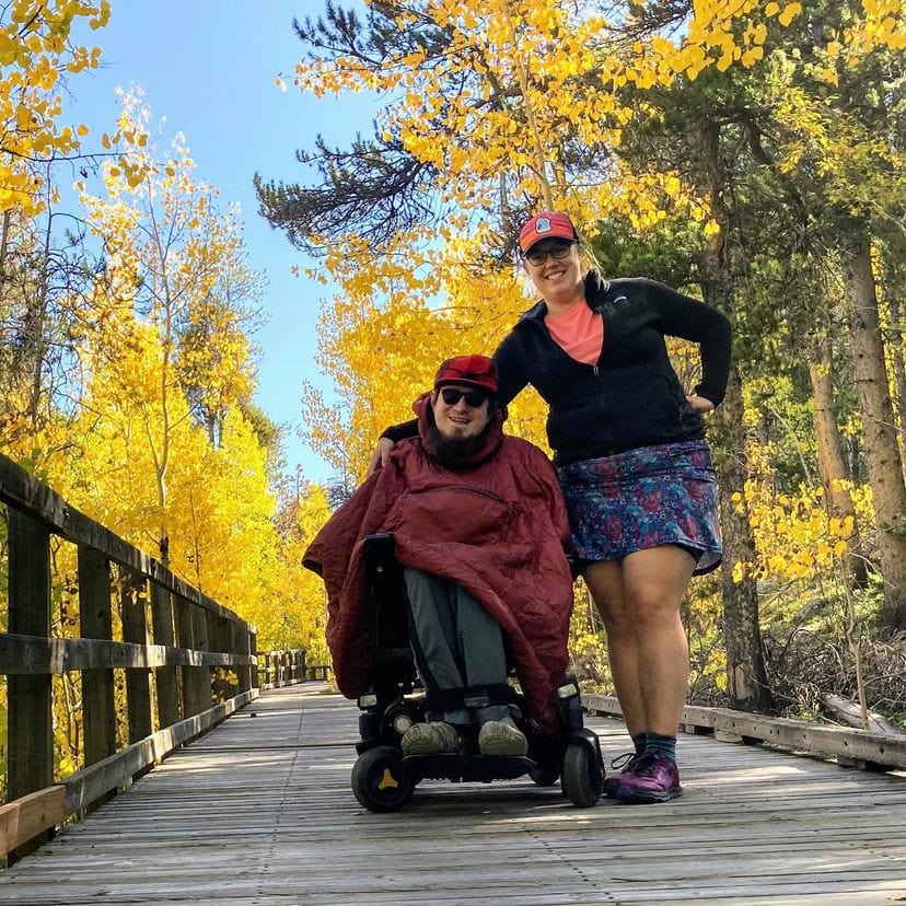 woman standing next to man in powerchari on wooden bridge