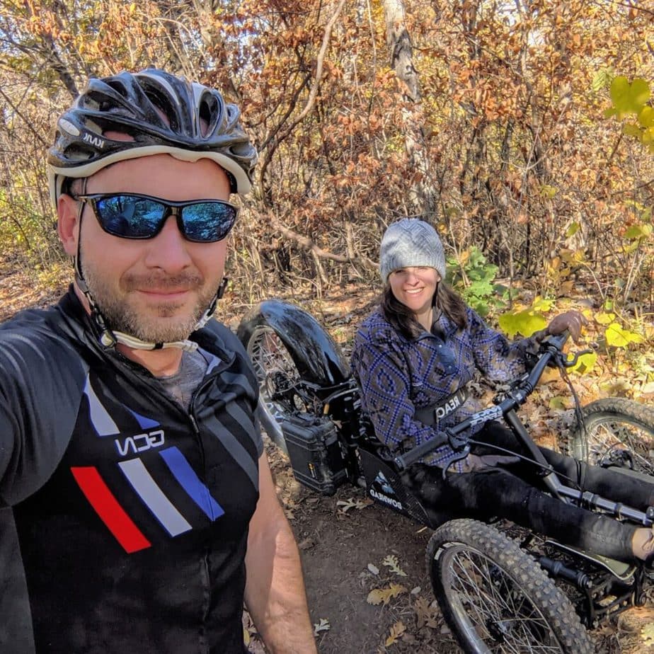 Shows woman on three-wheeled mountain bike and man with helmet and sunglasses, both smiling into camera. 