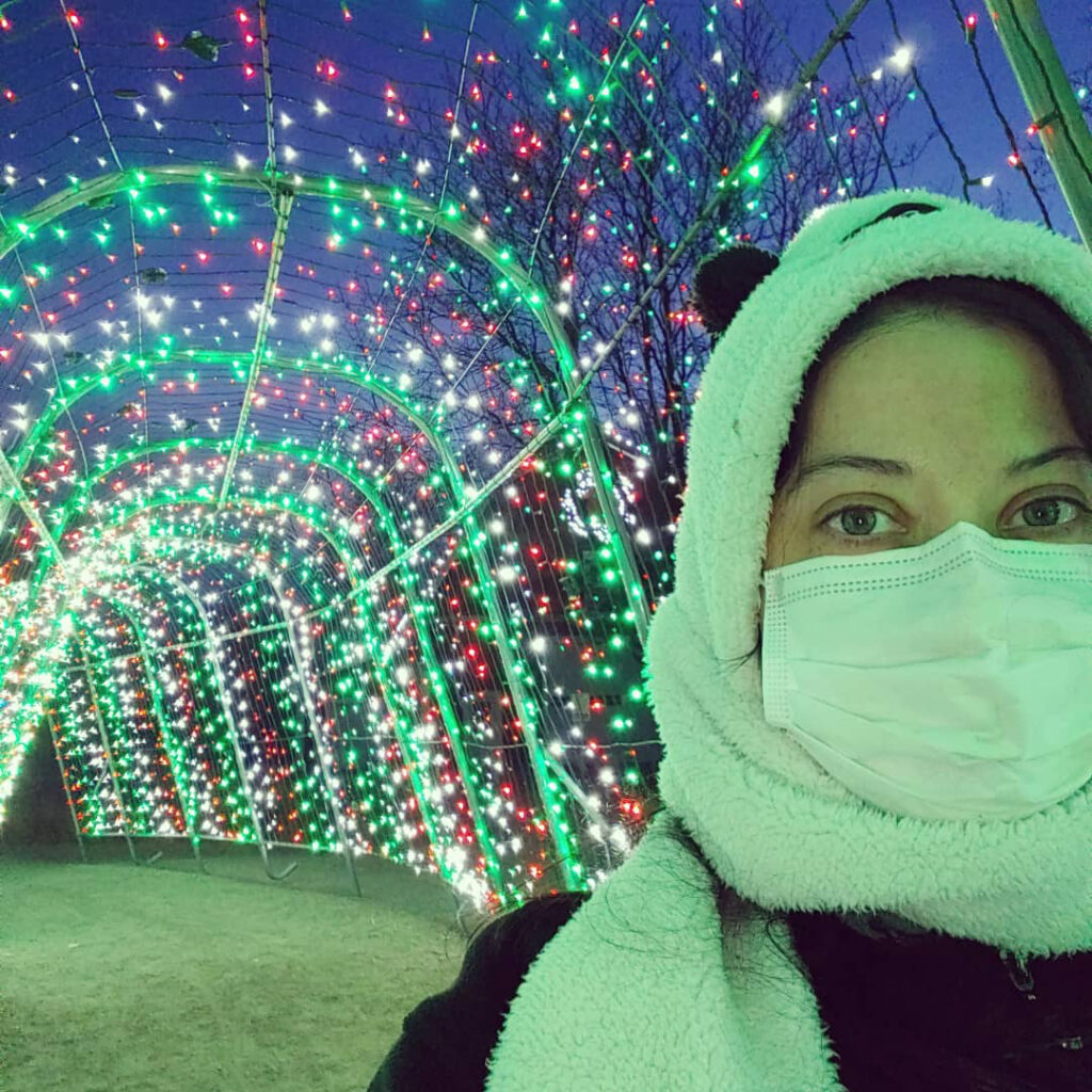 headshot of woman wearing hat, scarf and mask in from fo Christmas light display