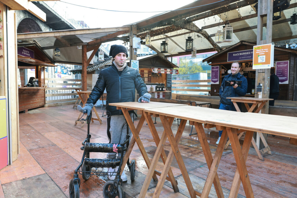 A man, Michel Rocatti wearing a winter coat and hat stands at a bar, with one hand on a walker and the other on a high table. He looks at the camera. 