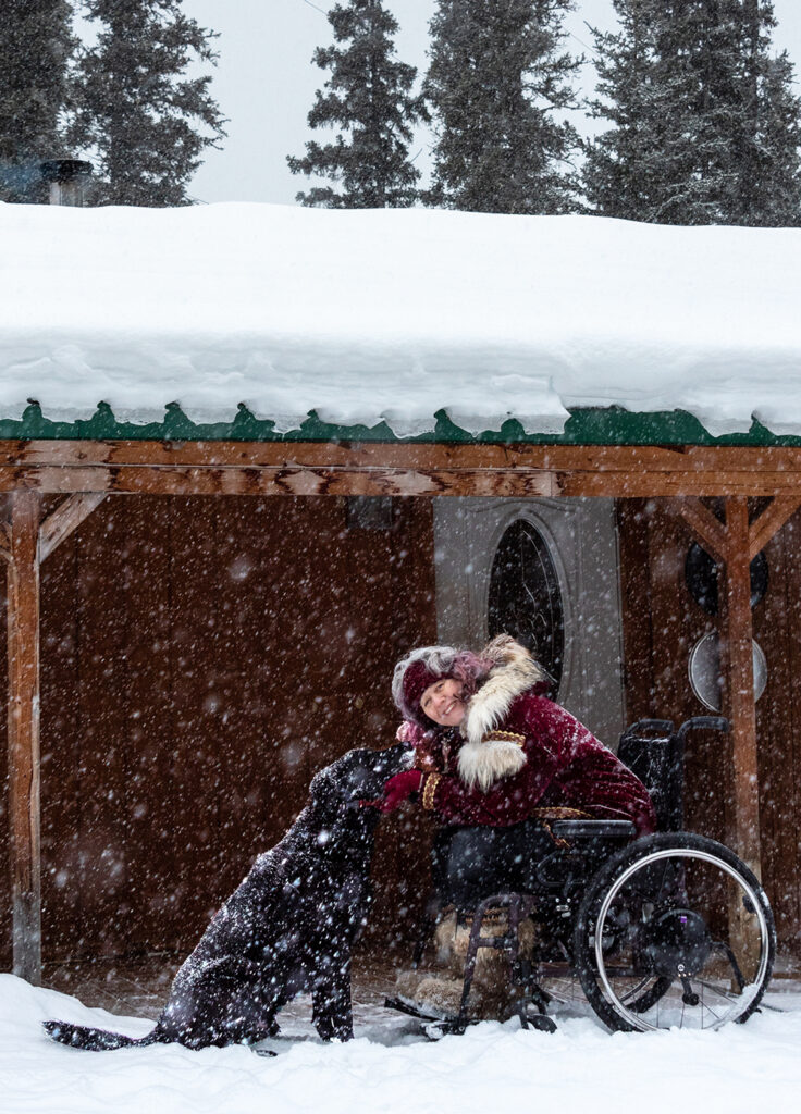 woman in wheelchair outside as snow is falling petting black dog