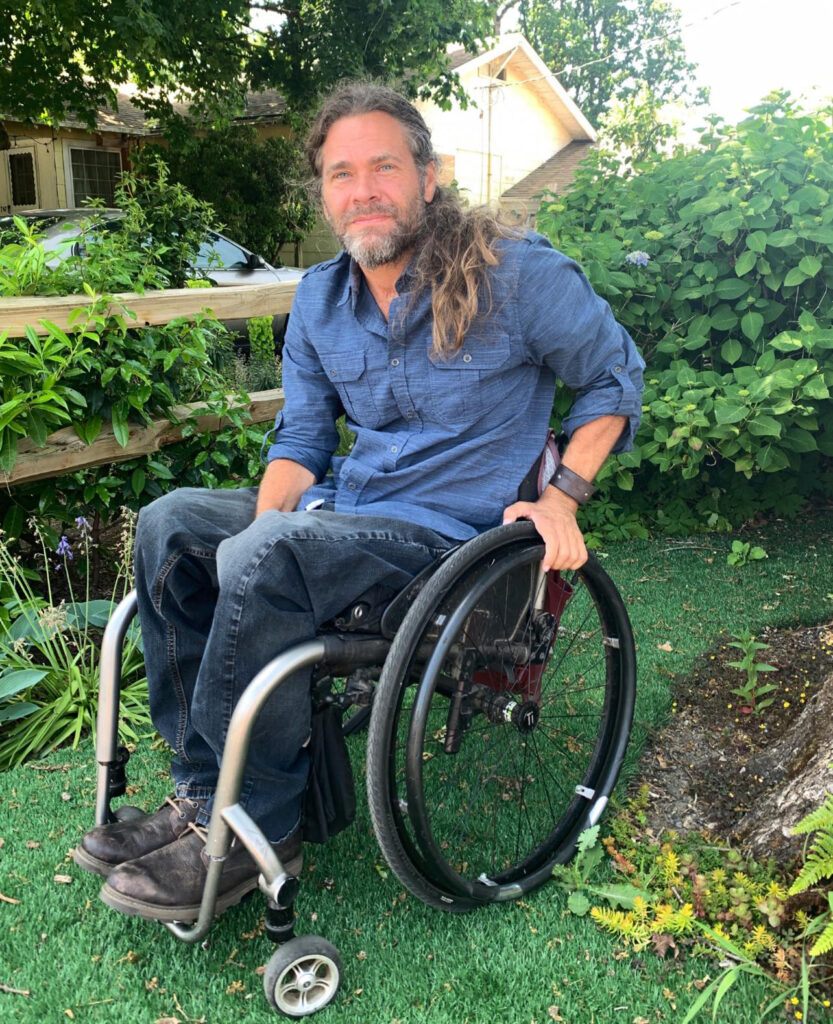 man in wheelchair outdoors, bushes and wood fence in background