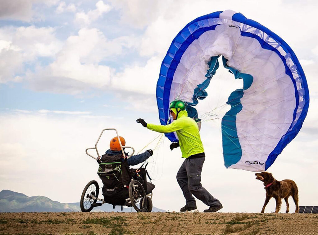man assisting man in paraglider with chute while dog watches