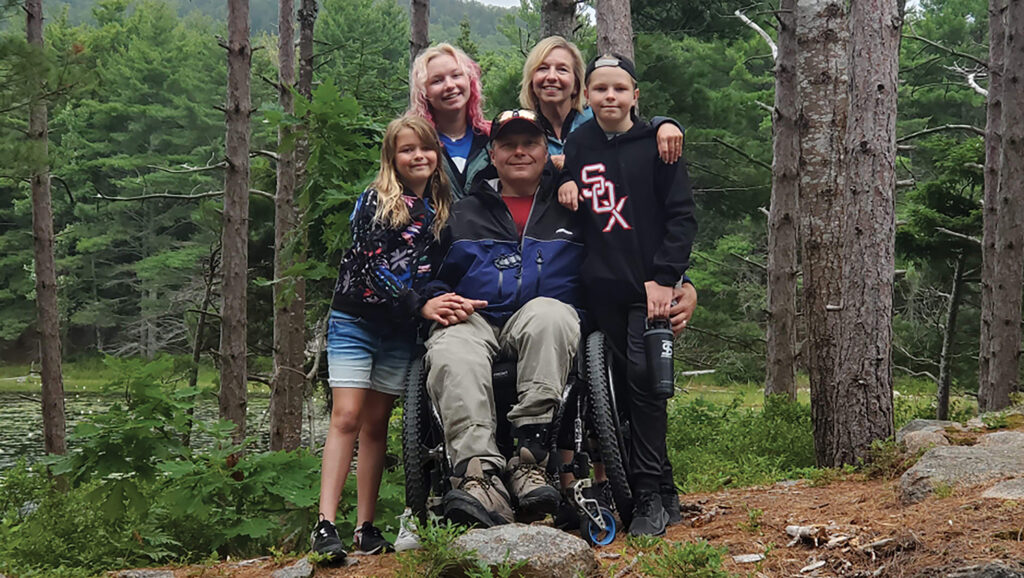 man in wheelchair surrounded by wife, son and two daughters in park woods setting