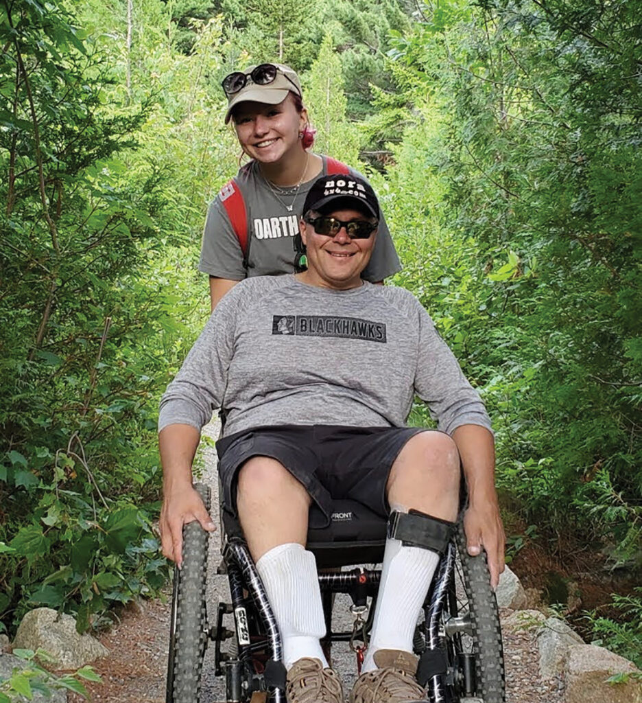 man in all terrain chair being pushed by daughter through wooded trail