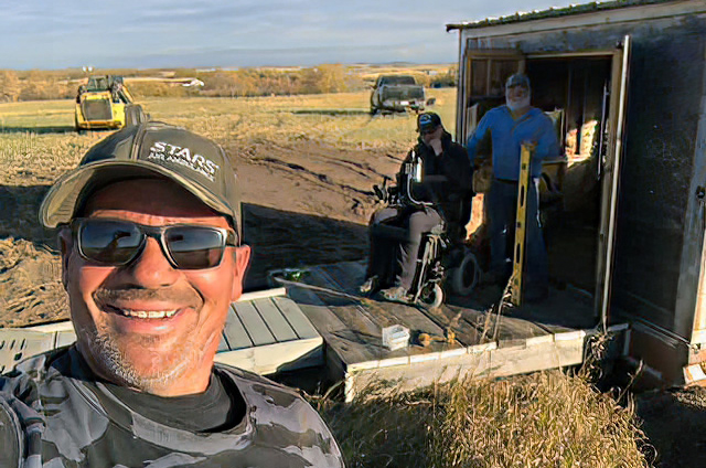 man taking selfie with two friends in background - one in a powerchair - in front of a hunting shack