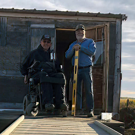 Author in powerchair with friend at top of wooden ramp in front of hunting shack 