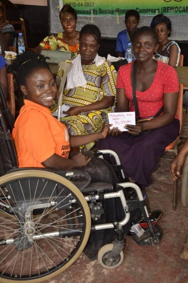 Black woman in orange shirt sitting in manual wheechair smiles at camera while handing an envelope to two other women.