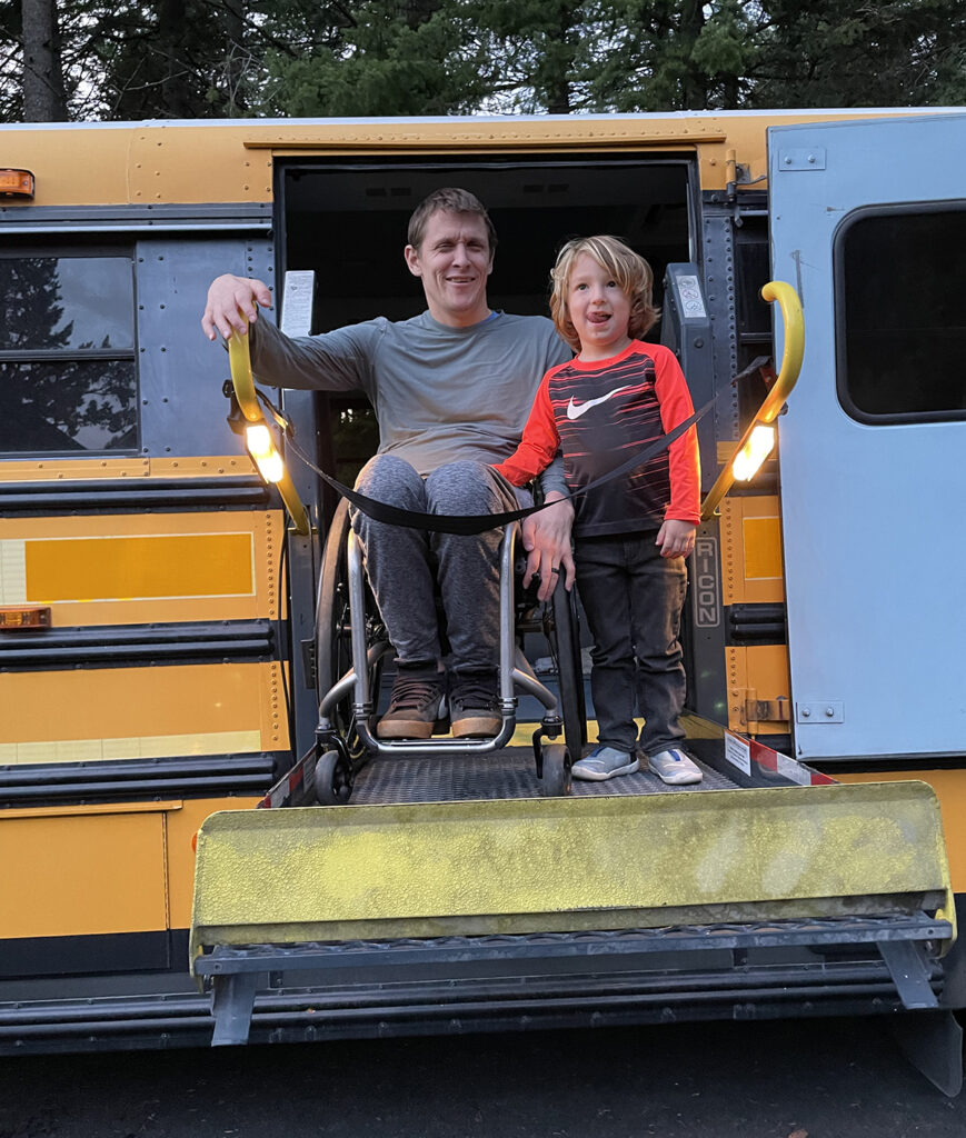 young child standing next to man in wheelchair on a lift on the side of a bus