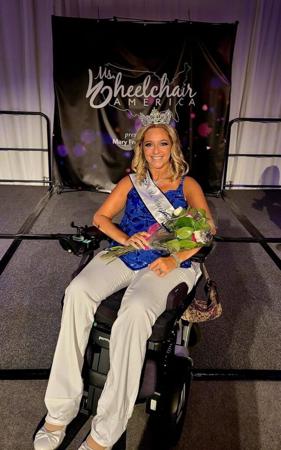 blond woman using power wheelchair wearing sash and crown and holding flowers, smiling at camera.
