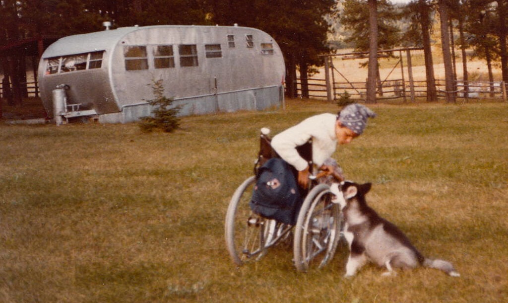 Old photo of woman in a field sitting in wheelchair leaning down to pet a dog, old mobile home in background