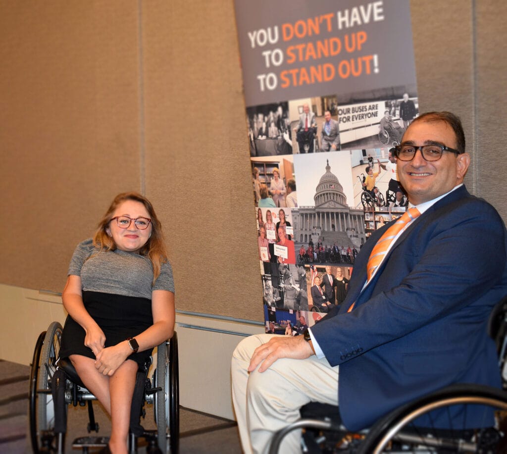 man and woman in wheelchairs in front of poster that reads: You don't have to stand up to stand out!