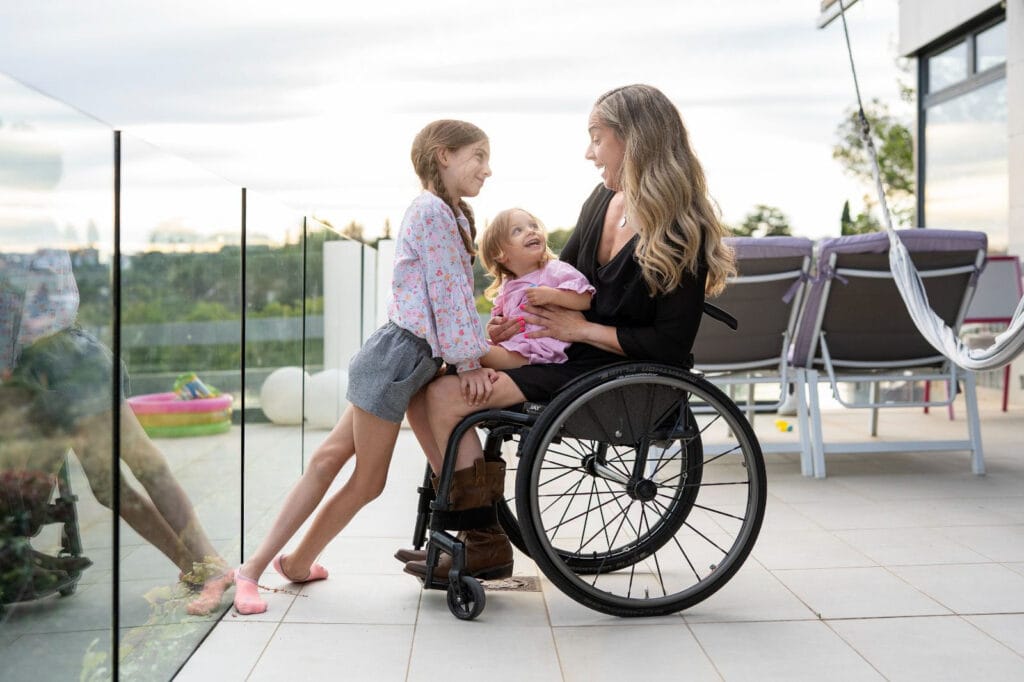 Woman in manual wheelchair wearing black dress playing with two young girls on patio.