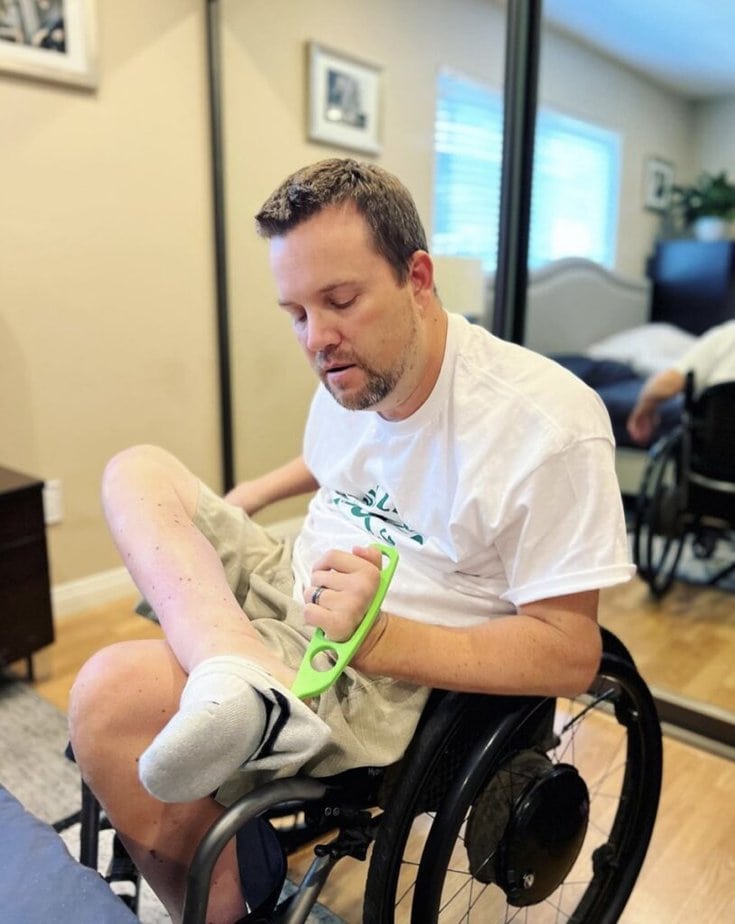 Man in wheelchair sitting with one leg crossed over the other, using a green hook to pull on his sock.