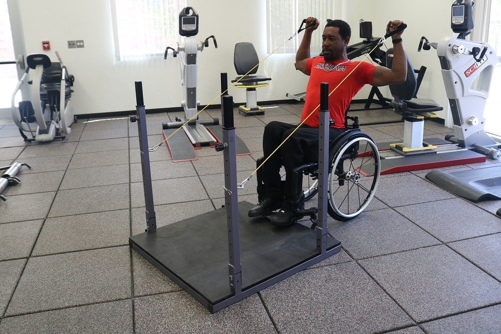 Man in manual wheelchair externally rotating shoulders holding workout bands in each hand. Bands are attached to metal frame of home gym. 
