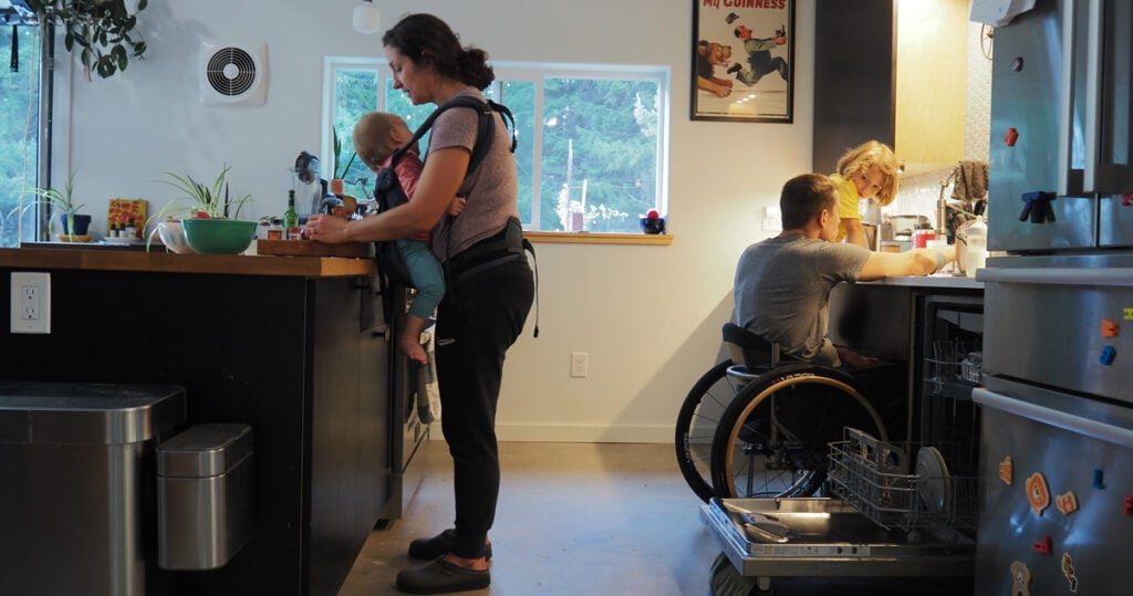  woman standing at kitchen counter with baby strapped on front, man in wheelchair behind her working with young child on back counter