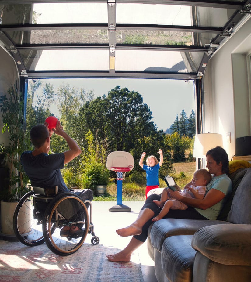 man in wheelchair in living room throwing toy basketball to young child outside, separated by open glass garage door. Woman and baby seated on couch