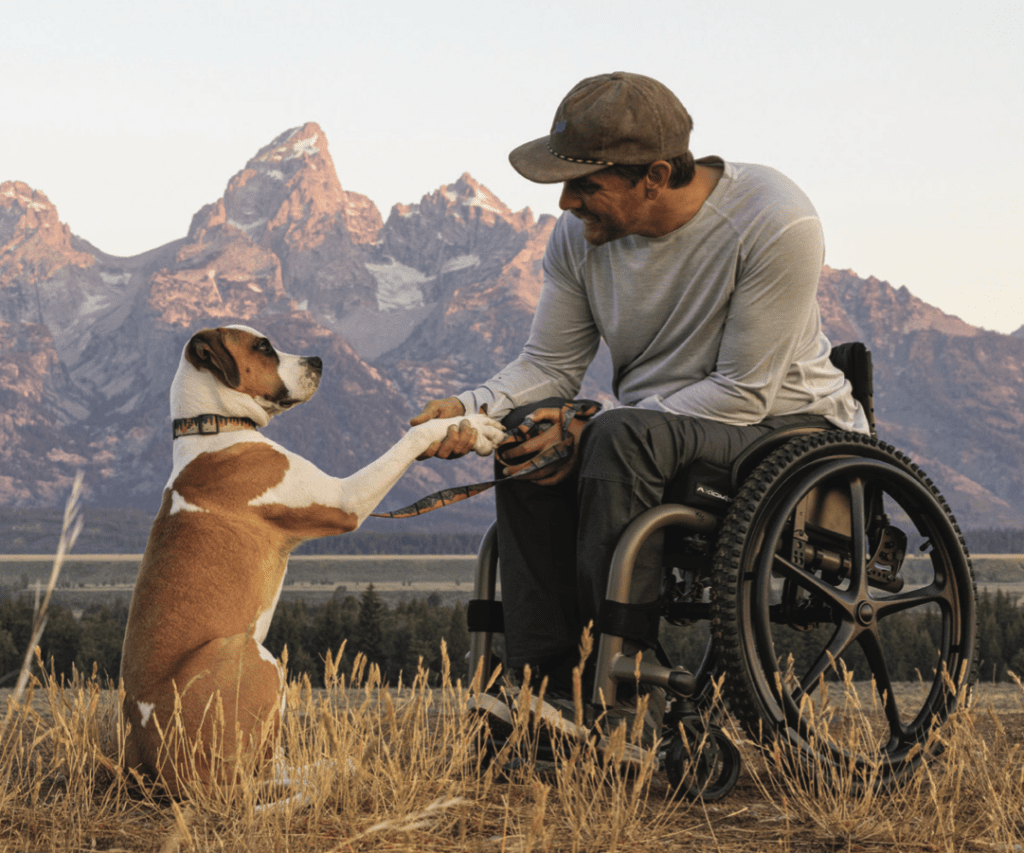 Homem sentado em cadeira de rodas manual em campo, sacudindo a pata do cachorro.  Montanhas ao fundo.