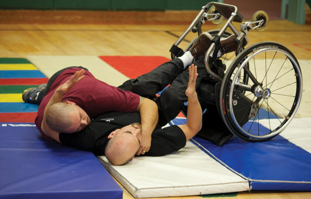 man in wheelchair doing martial arts with man on mat