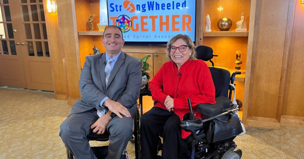 man in wheelchair wearing suit along side woman in powerchair with red shirt in front of StrongWheeled Together logo displayed on tv