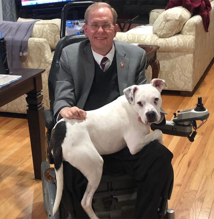 man in powerchair smiling at camera with white dog in his lap