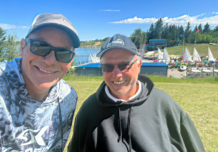 Two men taking a selfie with sailboats  in marina and lake in background