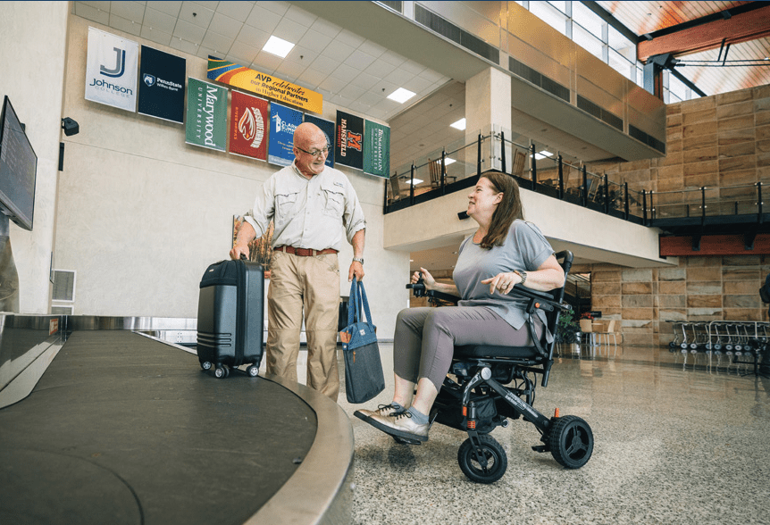 Woman and man at airport baggage carousel. Man is picking up bags and woman is sitting in Jazzy Carbon wheelchair and smiling.