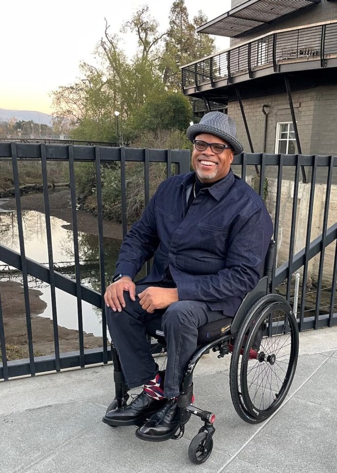 Black man sitting in wheelchair on bridge over a creek. He is wearing a collared jacket and a Fedora hat