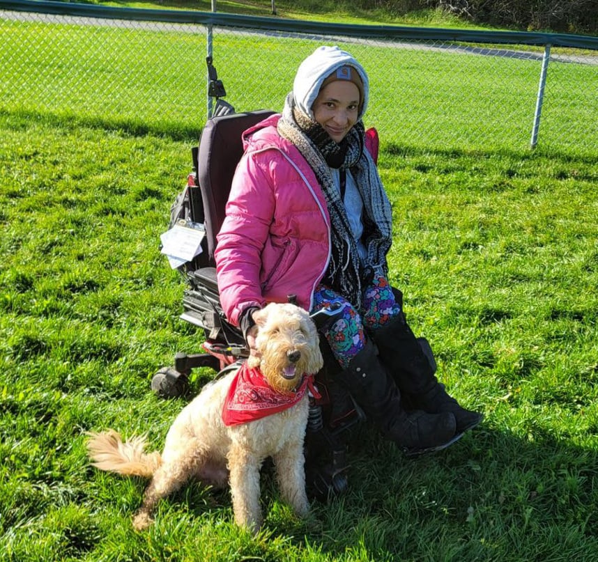 woman in powerchair outdoors wearing hat and coat, dog by her side