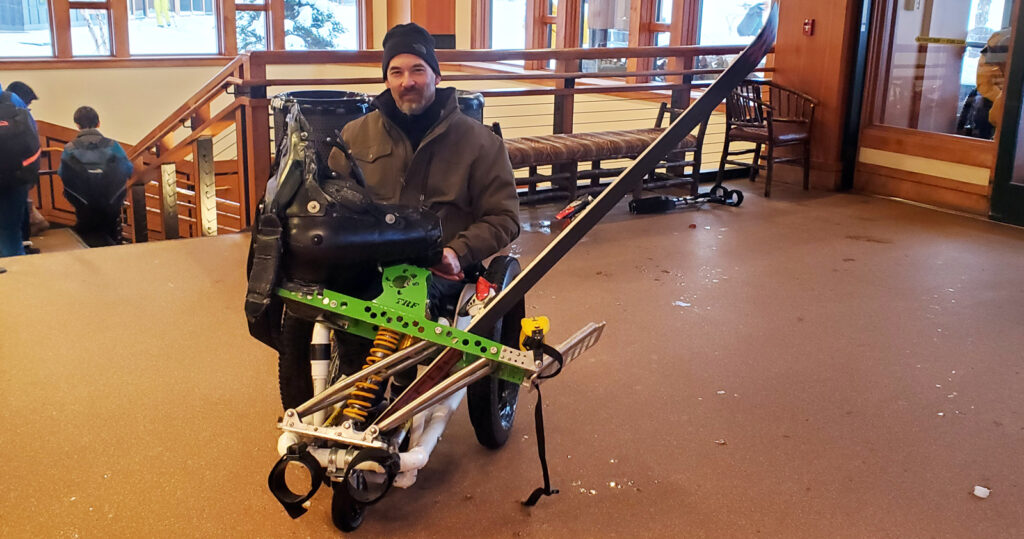 man in PVC constructed wheelchair transporting a ski and equipment