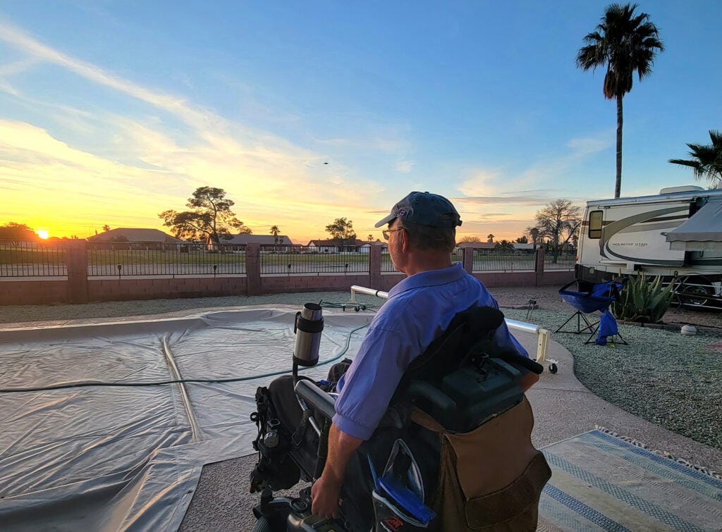 man shown from behind, sitting in powerchair watching the sunset by a covered pool