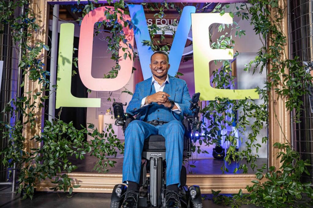 Black man sitting in power wheelchair, wearing suit and smiling at camera, he in front of hanging signs that spell out LOVE.