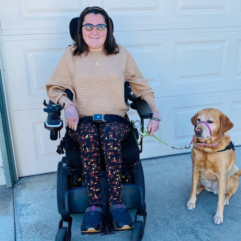 White woman sitting in power wheelchair wearing sunglasses. She holds a leash and a yellow lab sits by her side. 