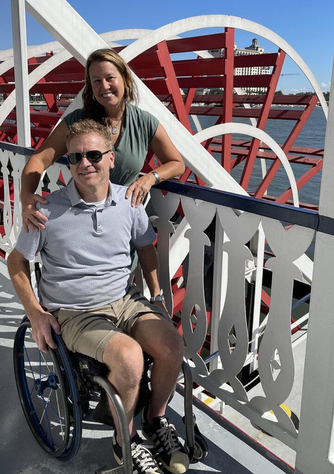 Woman standing next to man in wheelchair aboard a riverboat with paddle in background